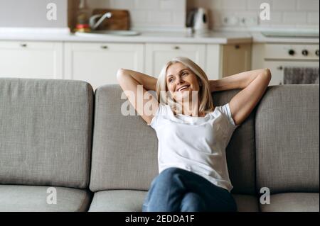 Glücklich reifen kaukasischen Frau ist auf der Couch im Wohnzimmer zu Hause entspannen. Frau mittleren Alters genießen Wochenende oder Freizeit von Hausaufgaben sitzen auf dem Sofa, schaut weg und lächelnd Stockfoto