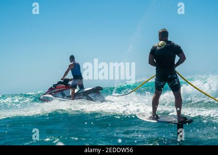 Ein Mann auf einem Tragflächenboot Surfbrett wird von einem Mann auf einem Jetski in einem türkisfarbenen Meer, klaren blauen Himmel, Sommertag geschleppt. Stockfoto