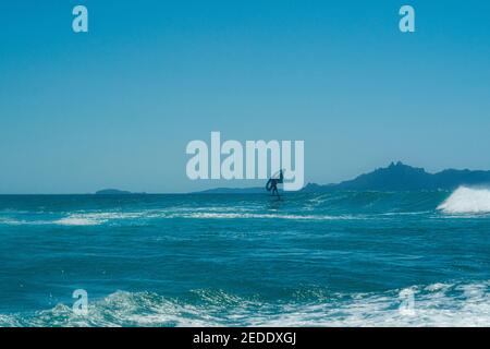 Ein Mann Flügel Folien mit einem Tragflügelboot Surfbrett und Hand gehalten aufblasbaren Flügel, auf einer türkisfarbenen Welle mit einem hellen blauen Himmel und einer Insel in der Ferne. Stockfoto