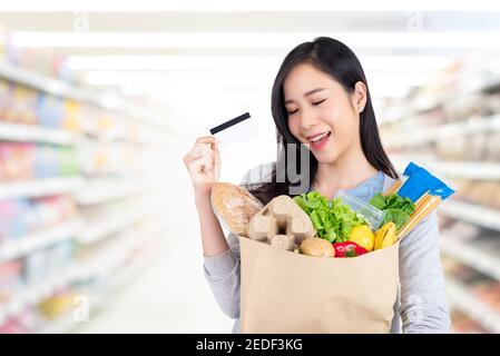 Schöne asiatische Frau im Supermarkt einkaufen mit Kreditkarte und Papiertüte voller Lebensmittel aufbewahren Stockfoto