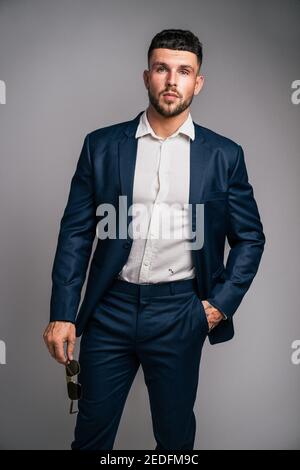 Porträt eines elegant gekleideten ernsthaften Mannes in Anzug, mit Sonnenbrille in der Hand, während er in die Kamera schaut. Isoliert auf grauem Hintergrund. Stockfoto