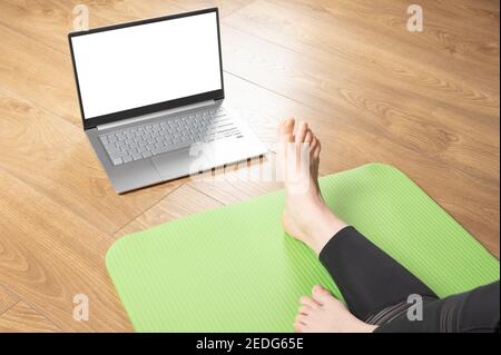 Frau sitzt auf Yoga-Matte mit ihren Beinen ausgestreckt und schaut in mockup von Laptop mit leeren weißen Bildschirm. Frau mit Laptop-Computer Training bei l Stockfoto