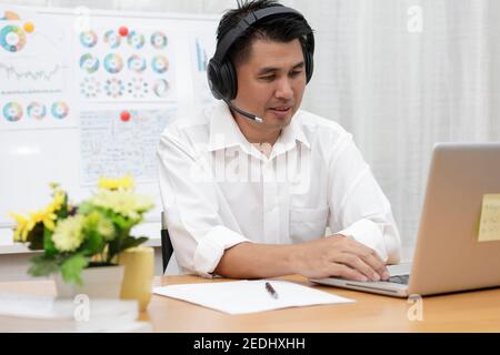 Asiatische Geschäftsmann im Gespräch mit Team über Videokonferenz schriftlich Notiz auf Papier. Geschäftsleute mit Laptop und Headset für Online-Meeting. Mann mit Stockfoto