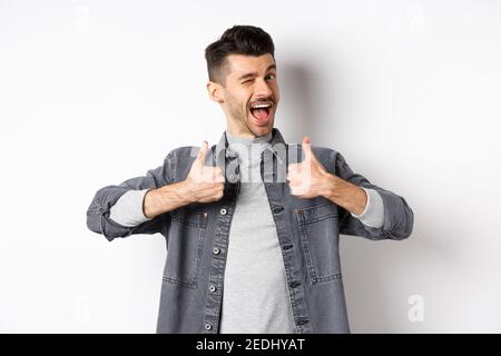 Glücklicher junger Mann mit Schnurrbart winkend und zeigt Daumen-up, empfehlen etwas, Lob gute Sache, Kompliment, stehend auf weiß Stockfoto