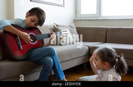 Kleiner hübscher Junge, der Gitarre spielt. Hübscher kleiner Junge, der Gitarre spielt, um sein Publikum zu Hause. Um seine Mutter, Schwester und Cousine. Stockfoto