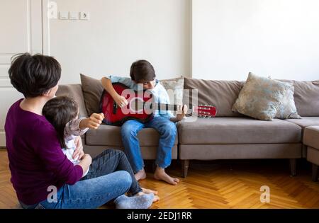 Kleiner hübscher Junge, der Gitarre spielt. Hübscher kleiner Junge, der Gitarre spielt, um sein Publikum zu Hause. Um seine Mutter, Schwester und Cousine. Stockfoto