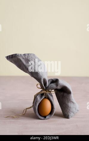 Kreativer Osterhase, naturbelassenes, unbemaltes Hühnereiei ist in eine graue Serviette gehüllt und steht auf beigem Hintergrund. Minimalismus, braun monochrom, verti Stockfoto