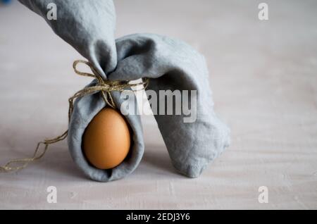 Kreativer Osterhase, naturbelassenes, unbemaltes Hühnereiei ist in eine graue Serviette gehüllt und steht auf beigem Hintergrund. Minimalismus, braun monochrom, verti Stockfoto