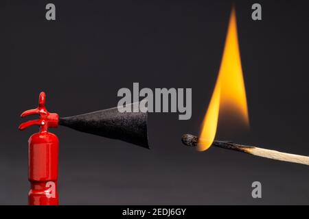 Ein winziger Spielzeugfeuerlöscher neben einem brennenden Streichholz vor einem beleuchteten schwarzen Hintergrund. Makroaufnahme, geringe Schärfentiefe. Stockfoto