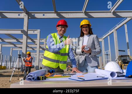 Glückliche weibliche Architektin und Geschäftsmann mit Daumen nach oben Blick auf Kamera. Mann und Frau in Schutzkleidung stehen neben dem Tisch mit Blueprint. Stockfoto