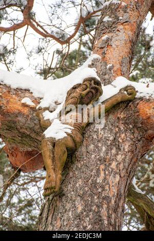 Schöne Holzskulptur von Christus oder Jesus von Schnee bedeckt hängen aus dem Stamm eines Baumes in einem Wald oder Park im Winter, vertikal Stockfoto