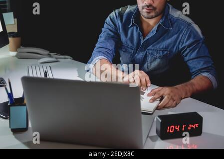 Workaholic Mann, der Überstunden spät in der Nacht im Büro bleibt Arbeiten an seinem Schreibtisch und Blick auf Laptop-Computer Stockfoto