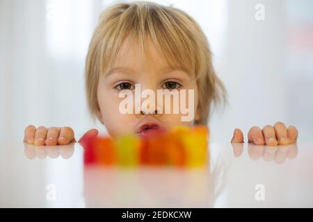 Cute Kleinkind blonde junge, Blick auf bunte Gummibärchen, sitzen auf dem Tisch Stockfoto