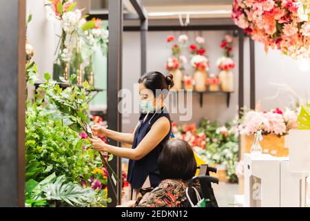 Ältere Frau mit Tochter in Schutzmaske wählt künstliche Blumenzweig im Geschäft. Stockfoto