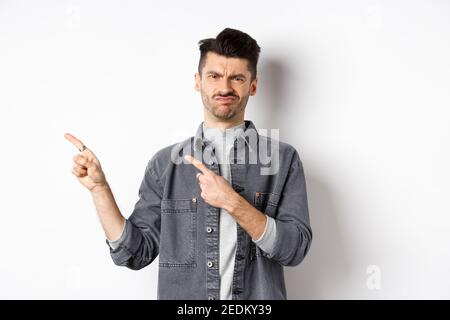 Skeptisch und enttäuscht junger Mann zeigt Finger nach links, Grimacing und Schütteln Kopf in Ablehnung, nicht wie schlechtes Produkt, auf weißem Hintergrund stehen Stockfoto