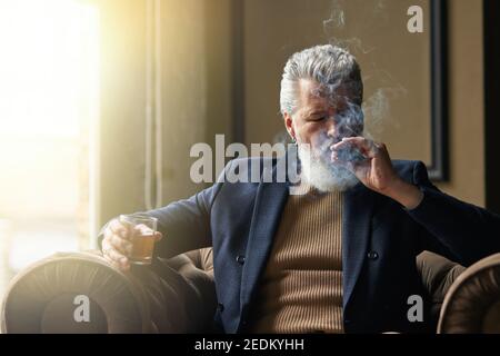 Porträt eines eleganten, reifen Geschäftsmannes, der Zigarre raucht und Whisky trinkt, während er sich in einem Stuhl im Loft-Interieur entspannt. Lifestyle, Erfolg, Menschen Konzept Stockfoto