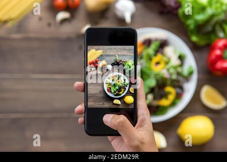 Food-Blogger mit Smartphone Foto von schönen Salat, rohe Spaghetti und Zutaten auf Holztisch zu teilen in den sozialen Medien Stockfoto