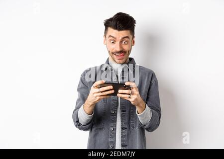 Aufgeregt Kerl spielen Videospiel auf Handy mit fokussierten Gesicht, tippen Sie auf Smartphone-Bildschirm, halten Handy horizontal, weißer Hintergrund Stockfoto