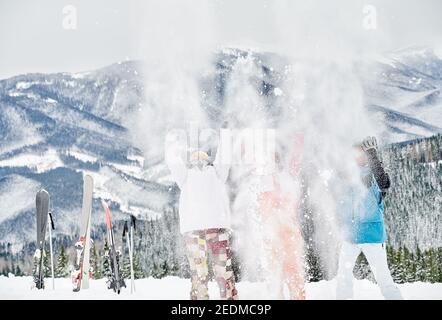 Drei Skifahrer in Skianzügen werfen frischen Pulverschnee hoch in die Luft, Spaß im Skigebiet mit schönen Bergen im Hintergrund. Konzept der Wintersport-Aktivitäten, Spaß und Beziehungen. Stockfoto