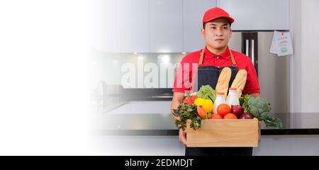 Lebensmittelgeschäft Lieferung Kurier Mann in roter Uniform und Schürze mit Lebensmittelkarton mit Lebensmitteln, frischem Obst und Gemüse. Liefert den Mann, der mit einer Holzkiste Lebensmittel umgeht Stockfoto