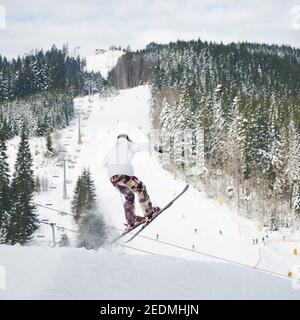 Männliche Skifahrer Skifahren in verschneiten Bergen mit verschneiten Bäumen und Hügeln im Hintergrund. Mann auf Skiern macht springen, während sie auf schneebedeckten Pisten gleiten. Konzept der aktiven Freizeit und Wintersport. Stockfoto