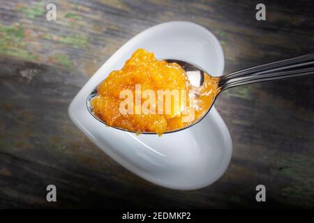 Löffel frische Orangenmarmelade . Unter ist weiße herzförmige Schüssel auf Holzhintergrund. Stockfoto