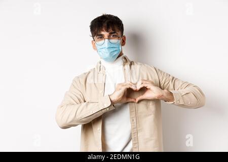 Health, covid und Quarantäne Konzept. Romantischer junger Mann in steriler medizinischer Maske mit Herzgeste auf der Brust, sag ich liebe dich, auf weiß stehend Stockfoto