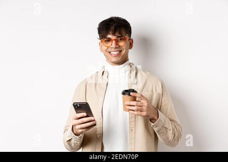 Natürliche schöner Mann in Gläsern trinken Kaffee aus Papiertasse und mit Handy, lächelnd zufrieden an der Kamera, weißer Hintergrund Stockfoto