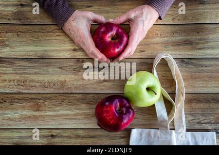 Hände einer alten Frau, die frischen Bio-Apfel hält. Eco Shopping Bag und rote und grüne Äpfel auf Holzhintergrund, Flat Lay Top View Stockfoto