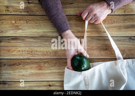 Hände einer alten Frau setzen frische Bio-Avocado in Eco-Einkaufstasche auf Holzhintergrund, Flat Lay. Draufsicht. Umweltschutzkonzept Stockfoto