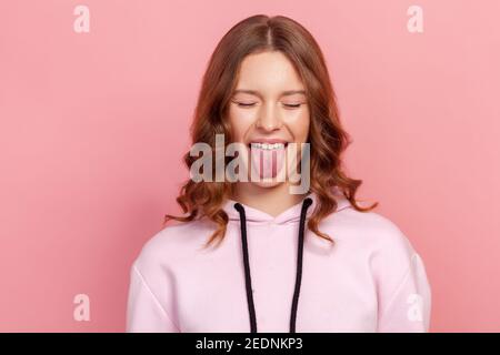 Portrait of adorable Teenager-Mädchen im Hoodie mit geschlossenen Augen zeigen Zunge, suchen verspielt frech, weiblich millenial Spaß. Innenaufnahme im Studio, Stockfoto