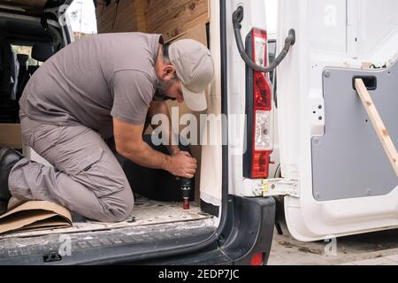 Detail eines Mannes, der mit einem Batteriewerkzeug ein Loch in den Boden eines Fahrzeugs bohrt. Selbstbau eines Wohnwagens auf der Straße. Stockfoto