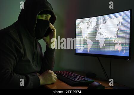 Maskierter Hacker, der auf seinem Telefon vor einem Computer anruft, um massive Angriffe auf Datenmissbrauch auf der ganzen Welt zu organisieren. Cyber-kriminelle Konzept. Stockfoto