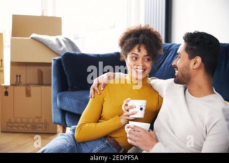 Junge gemischte ethnische Zugehörigkeit Paar, die eine Pause am bewegenden Tag In ein neues Zuhause auf dem Boden sitzend in der Lounge Kaffee trinken Umgeben von Umzugskartons