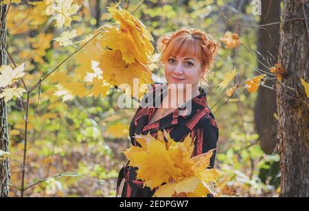 Fröhliche junge Frau steht im Herbstwald und hält Zwei große Blumensträuße aus gelben Blättern in ihm Stockfoto