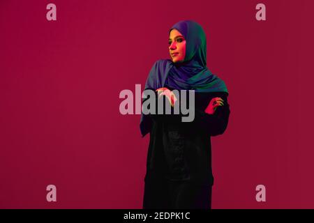 Jugendlich. Schöne arabische Frau in stilvollen Hijab isoliert auf burgunderrot Studio Hintergrund in Neonlicht mit Copyspace für Anzeige. Mode, Schönheit, Stilkonzept. Weibliche Modell mit trendigen Make-up, Accessoires. Stockfoto