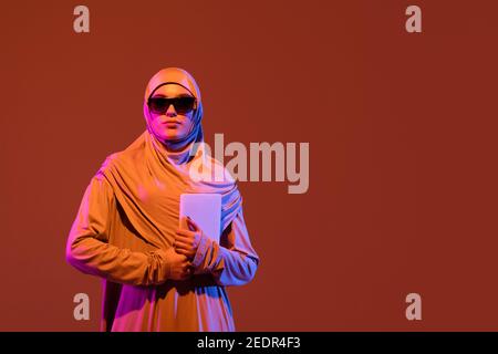 Schöne arabische Frau in stilvollen Hijab isoliert auf braunen Studio-Hintergrund in Neon, Copyspace für Anzeige. Mode, Schönheit, Stilkonzept. Weibliche Modell mit trendigen Make-up, Accessoires. Funktioniert mit Tablet. Stockfoto