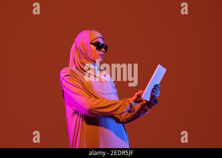 Schöne arabische Frau in stilvollen Hijab isoliert auf braunen Studio-Hintergrund in Neon, Copyspace für Anzeige. Mode, Schönheit, Stilkonzept. Weibliche Modell mit trendigen Make-up, Accessoires. Funktioniert mit Tablet. Stockfoto