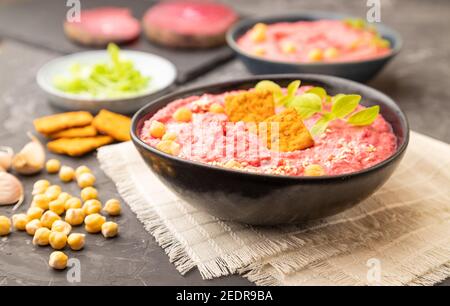 Hummus mit Rüben und mikrogrünen Basilikum Sprossen in Keramikschale auf schwarzem Beton Hintergrund und orangefarbenem Textil. Seitenansicht, Nahaufnahme, selektiver Fokus. Stockfoto