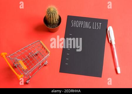 Einkaufsliste auf schwarzem Papier geschrieben. Konzept der Lebensmittel-Checkliste. Speicherplatz kopieren. Isoliert auf rotem Hintergrund. Neben der Liste befindet sich der Mini-Warenkorb. Stockfoto