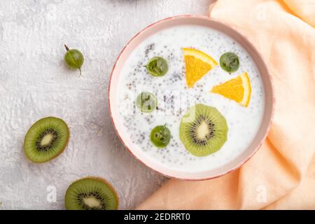 Joghurt mit Kiwi, Stachelbeere, Chia in Keramikschale auf grauem Betongrund und orangefarbenem Leinentextilien. Draufsicht, flach liegend, Nahaufnahme. Stockfoto