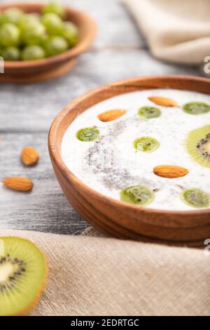 Joghurt mit Kiwi, Stachelbeere, Chia und Mandeln in Holzschüssel auf grauem Holzhintergrund und Leinentextilien. Seitenansicht, Nahaufnahme, selektiver Fokus. Stockfoto