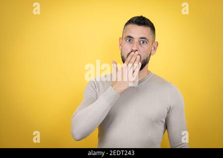 Schöner Mann mit Bart tragen Pullover über gelben Hintergrund bedeckt Mund mit der Hand, schockiert und Angst vor Fehler. Überrascht Ausdruck Stockfoto