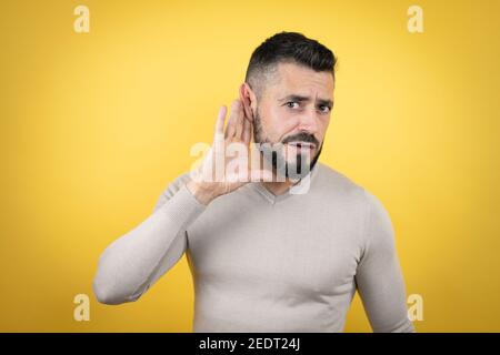 Schöner Mann mit Bart trägt Pullover über gelben Hintergrund überrascht Mit der Hand über das Ohr hören ein Gehör zu Gerücht oder Klatsch Stockfoto