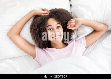 Glückliche junge afroamerikanische Frau, die im Bett liegt, versucht aufzuwachen, die Arme nach dem Schlaf im Schlafzimmer zu strecken Stockfoto