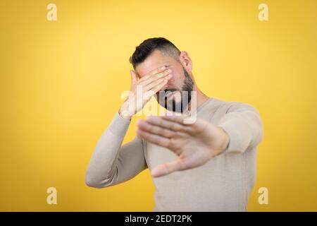 Schöner Mann mit Bart trägt Pullover über gelben Hintergrund Augen mit Händen und tun Stop-Geste mit traurig und Angst Ausdruck. Peinlich Stockfoto