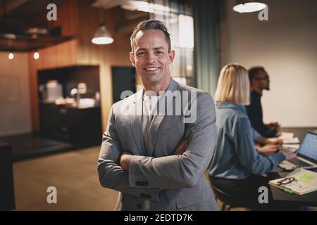 Lächelnder reifer Geschäftsmann, der mit gekreuzten Armen in einem steht Büro mit Kollegen, die an einem Tisch im Hintergrund sprechen Stockfoto