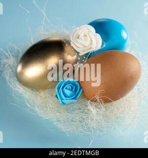 Baumaggs im Nest. Goldene, blaue und braune Eier im Nest. Frohe Ostern Konzept. Bemalte Eier in einem Nest mit einer blauen und weißen Rose auf einem pastellblauen Hintergrund Stockfoto