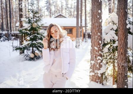 Winter sonnig kalt Porträt im Schneefall der schönen jungen Frau In einer weißen warmen outwear Daunenjacke im Hinterhof Landhaus Holzhaus auf dem Hintergrund der grünen Fichtenkiefer Bäume mit Schnee bedeckt Stockfoto
