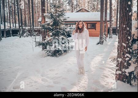 Winter sonnig kalt Porträt im Schneefall der schönen jungen Frau In einer weißen warmen outwear Daunenjacke im Hinterhof Landhaus Holzhaus auf dem Hintergrund der grünen Fichtenkiefer Bäume mit Schnee bedeckt Stockfoto